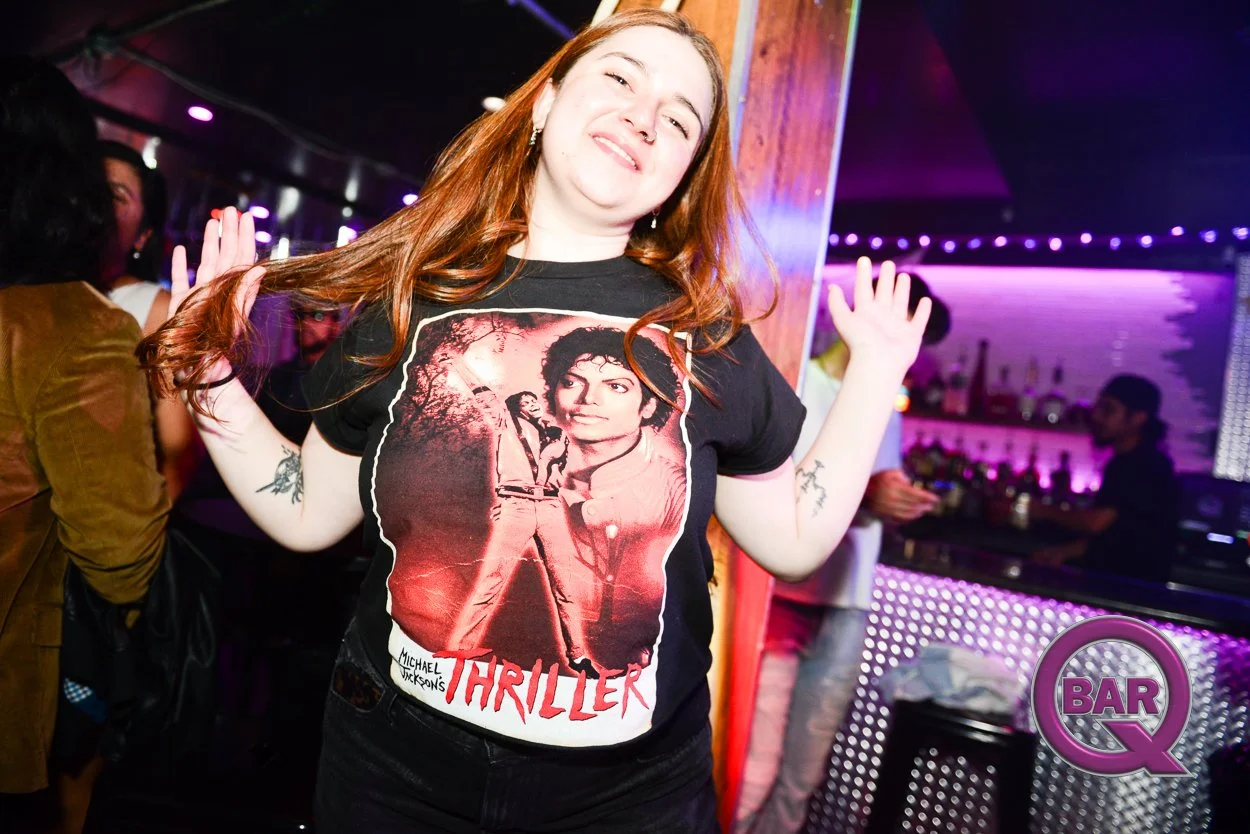 A young woman with long red hair dancing and smiling at a bar or nightclub. She is wearing a black T-shirt featuring Michael Jackson's Thriller album cover. The background shows other people, bar shelves, and purple nightclub lighting.
