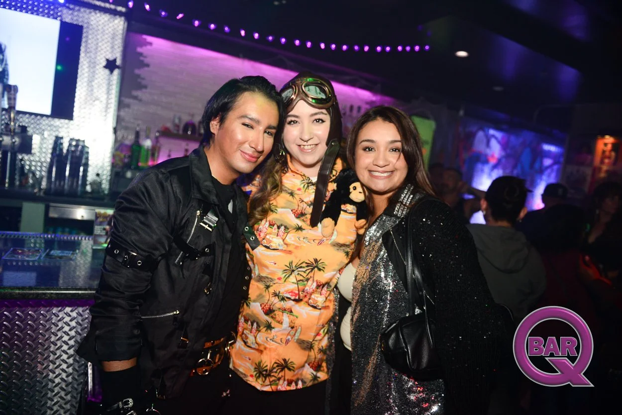 Three friends smiling at a bar with colorful lights, one wearing sunglasses and Hawaiian shirt, others in black and shiny jackets.