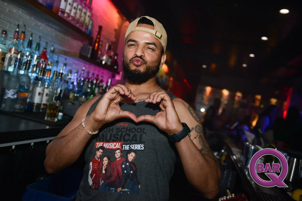 A man with a beard and tattoo on his arm making a heart shape with his hands at a bar, wearing a beige cap backwards, a sleeveless t-shirt with a "High School Musical" design, and a watch, in a dimly lit bar with bottles of alcohol on the shelves beh