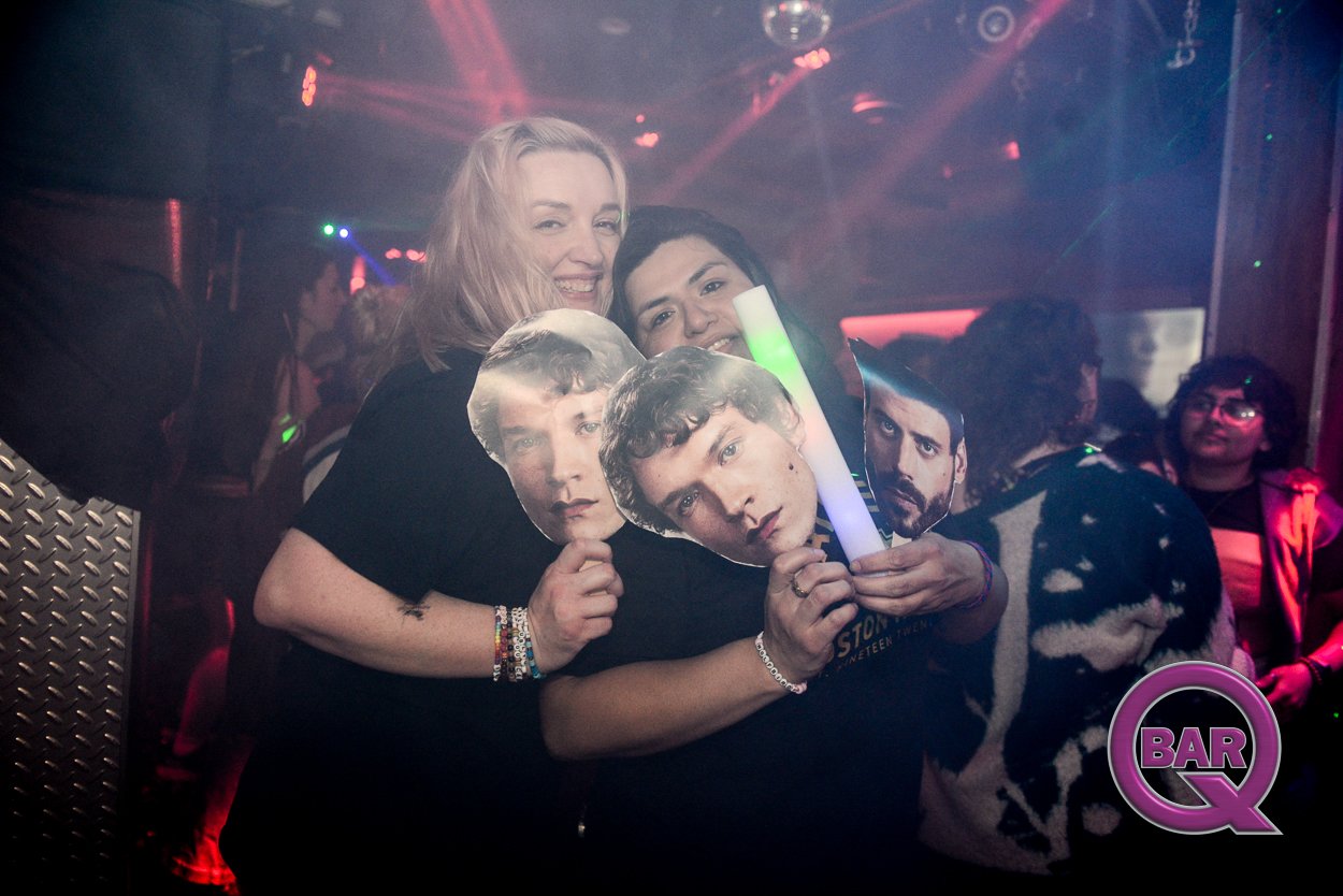 Two women celebrating at a nightclub holding cardboard cutouts of The Beatles members and a glowing light stick. The backdrop features colorful lights and other partygoers.