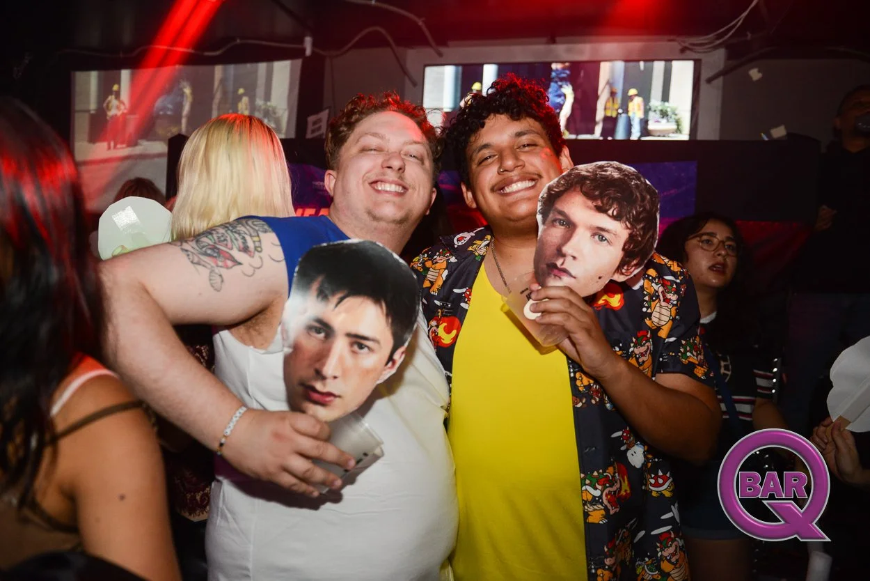 Two men at a bar holding cardboard cutouts of a man's face, smiling and enjoying themselves.