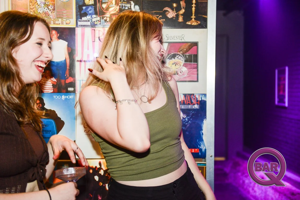 Two women laughing and talking at a bar, with posters on the wall behind them and purple lighting on the right.