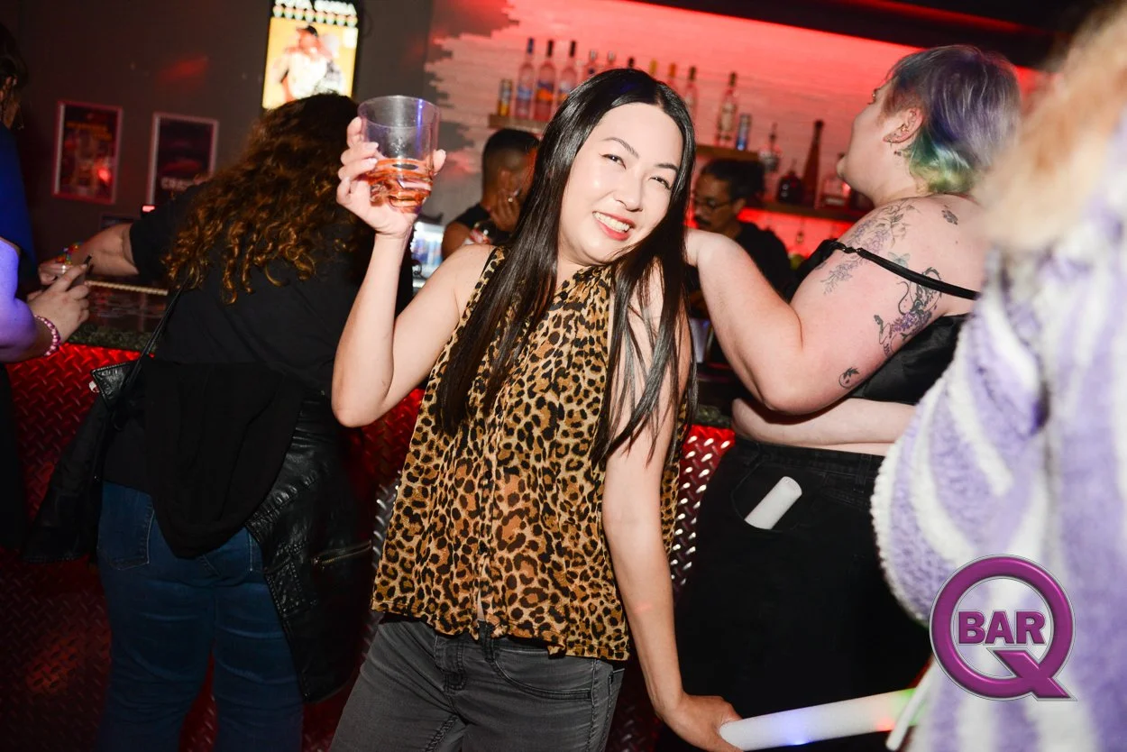 A woman in a leopard print top holding a drink, smiling, in a bar with people in the background.