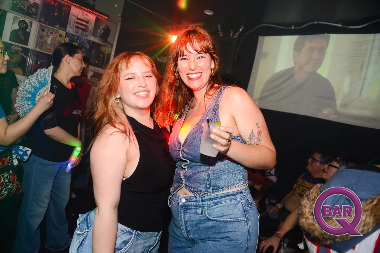 Two women smiling and posing for a photo in a bar or nightclub, with one holding a drink, surrounded by other people and colorful lights.