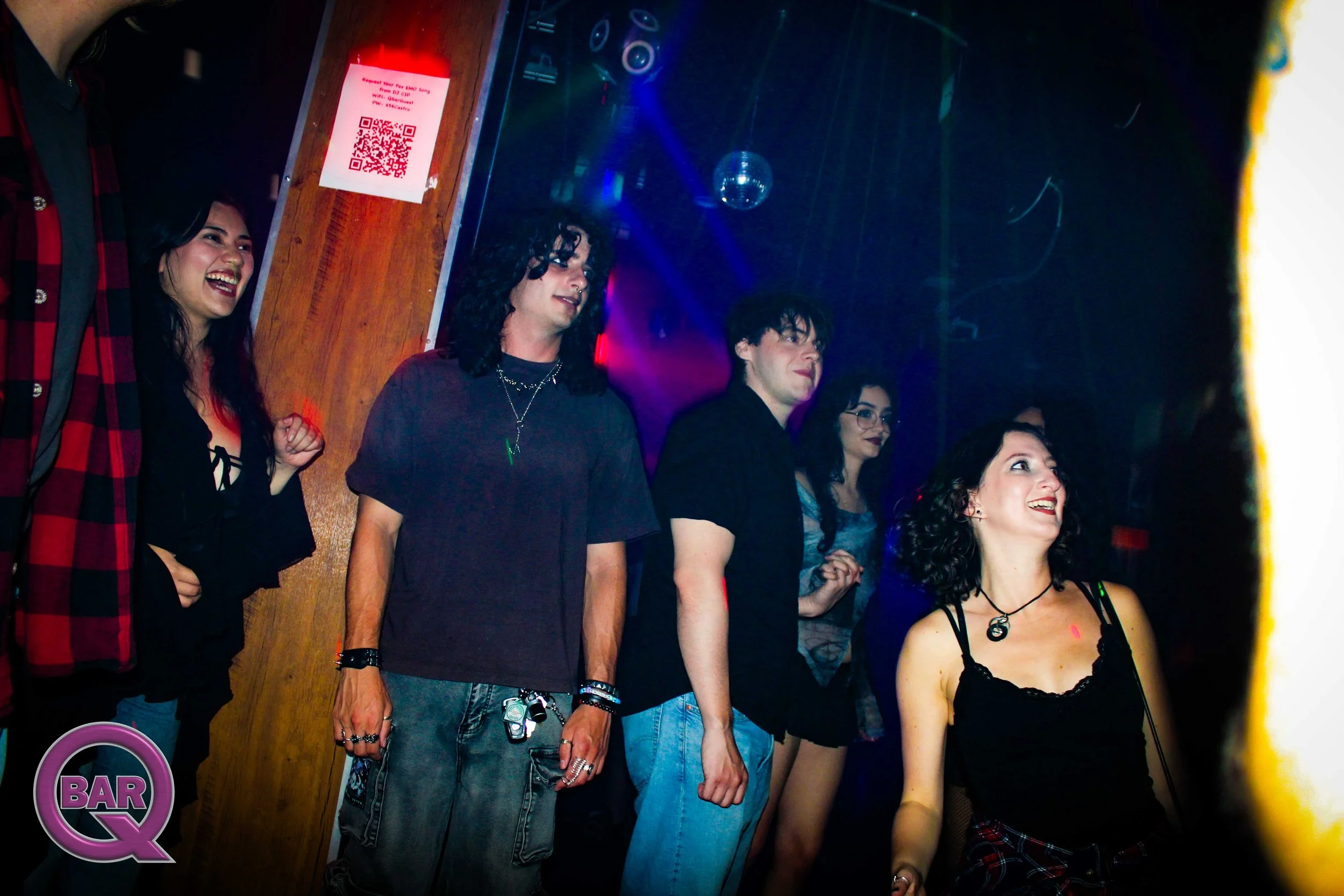 Group of five young adults in a dimly lit bar or nightclub, standing close together, smiling and engaging with each other.