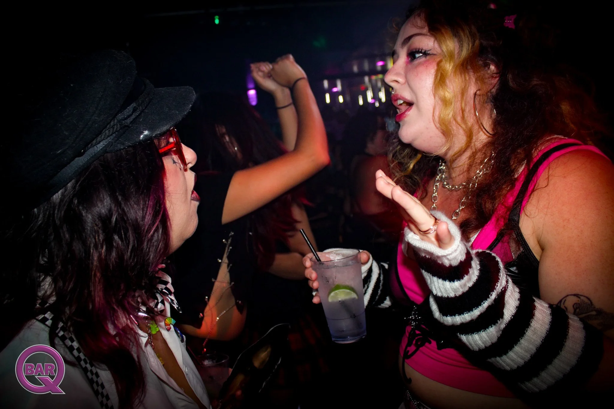 Two young women at a nightclub engage in conversation, one holding a drink with lime, the other wearing glasses and a witch hat.