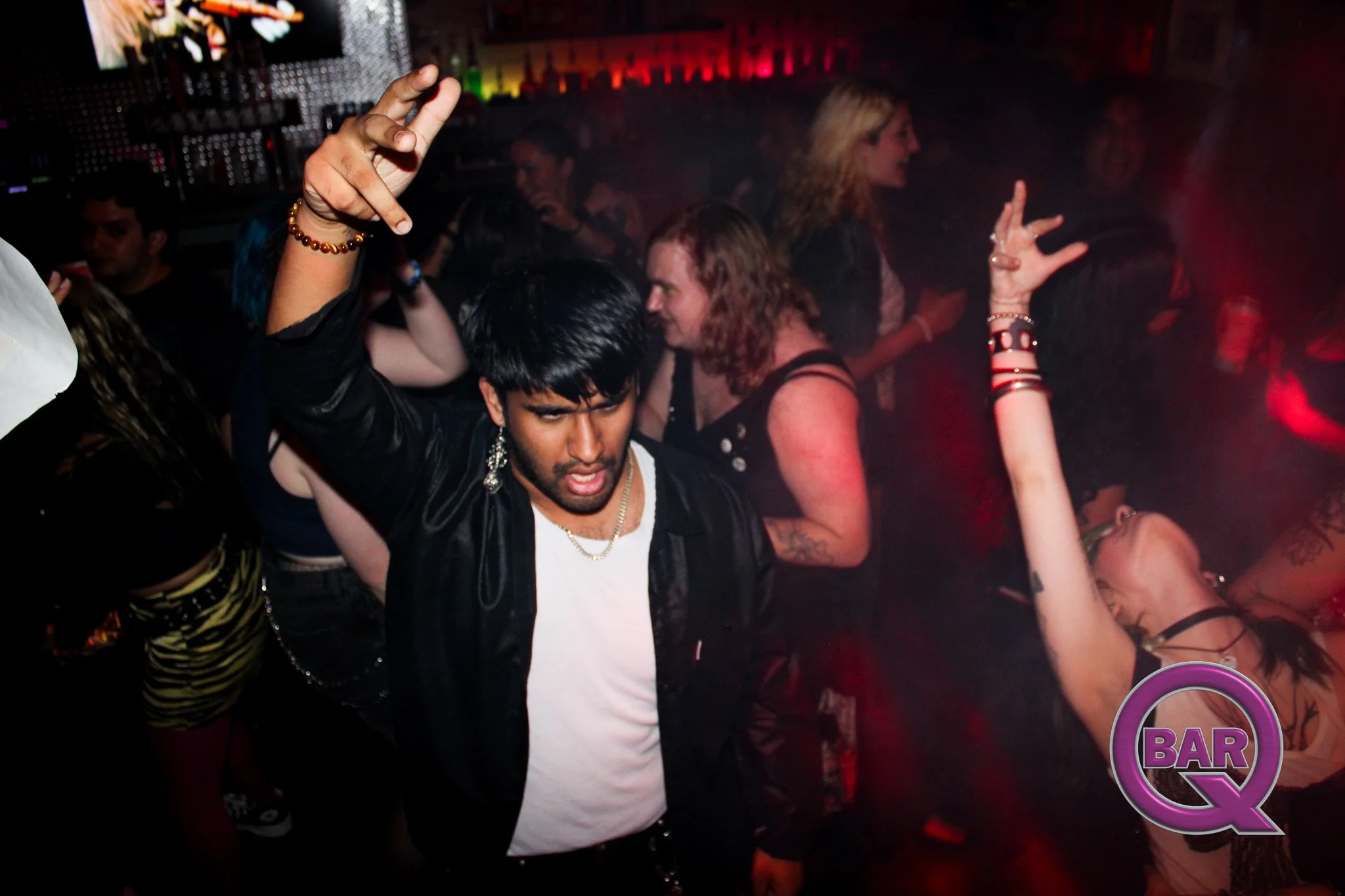 People dancing in a nightclub with colorful lights, one person in the foreground making a hand gesture.