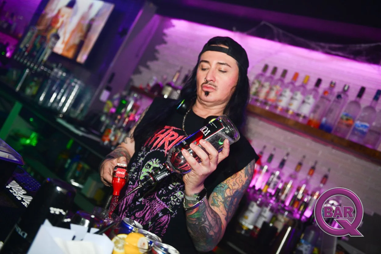A person with long dark hair, tattoos, and a black cap behind a bar counter, pouring liquor from a bottle into a glass, with bar equipment and bottles in the background.