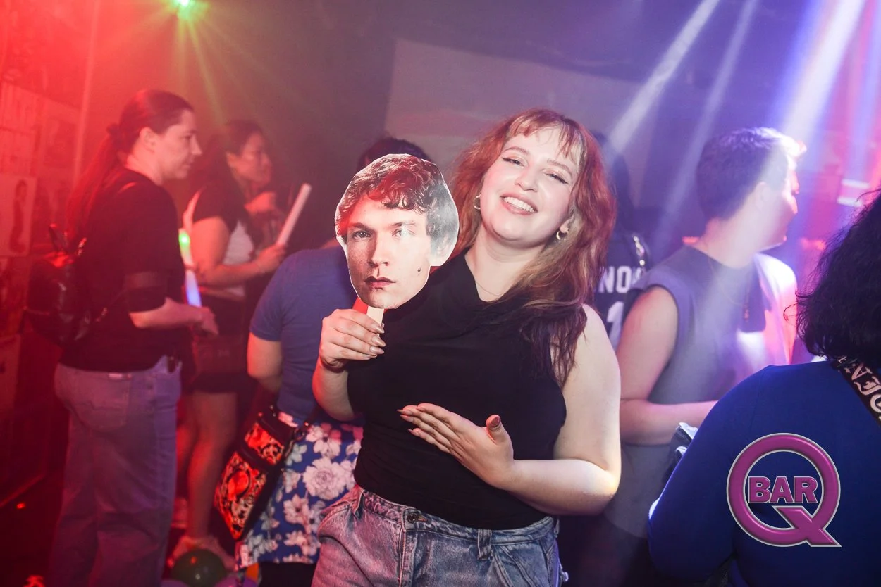 A woman smiling at a party, holding a cardboard cutout face of a man, surrounded by people in a dimly lit club with colorful lights.