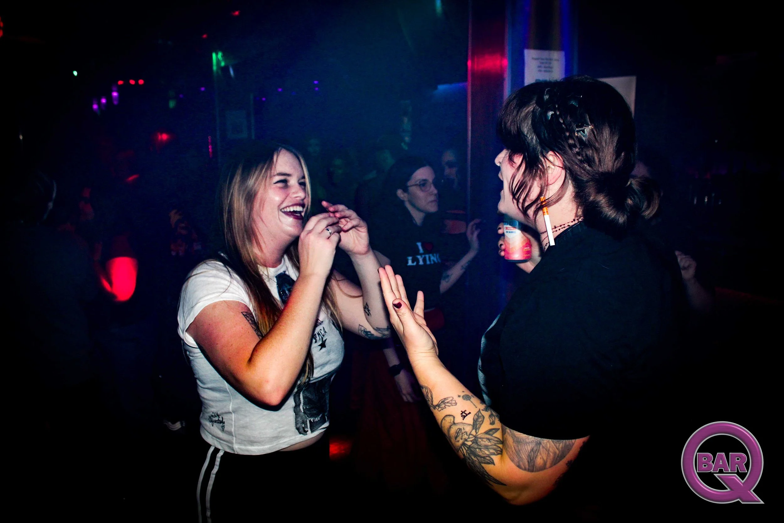 Two women are dancing and smiling at a nightclub with colorful lights. One has long blonde hair and tattoos on her arms, the other has dark hair styled in braids and is holding a drink.
