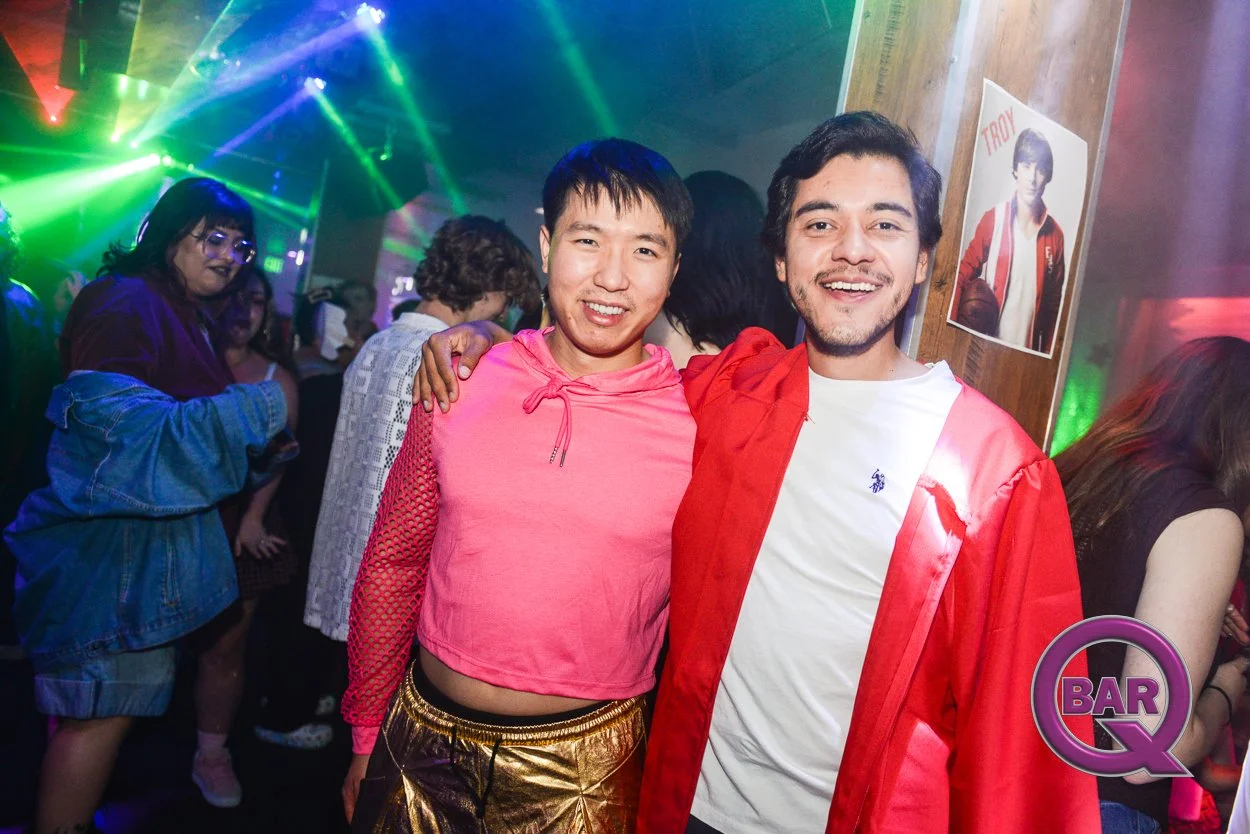 Two young men smiling at a party with colorful lights, one in a pink hoodie and gold pants, the other in a white t-shirt and red jacket, in a lively bar or nightclub.