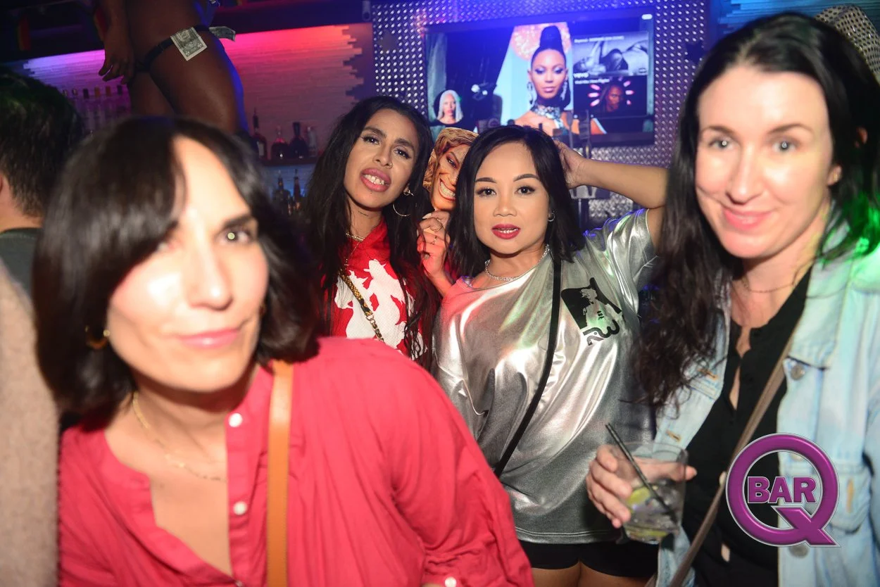Group of women at a bar or nightclub, some holding drinks, with colorful lighting and a large screen in the background displaying a woman’s portrait.