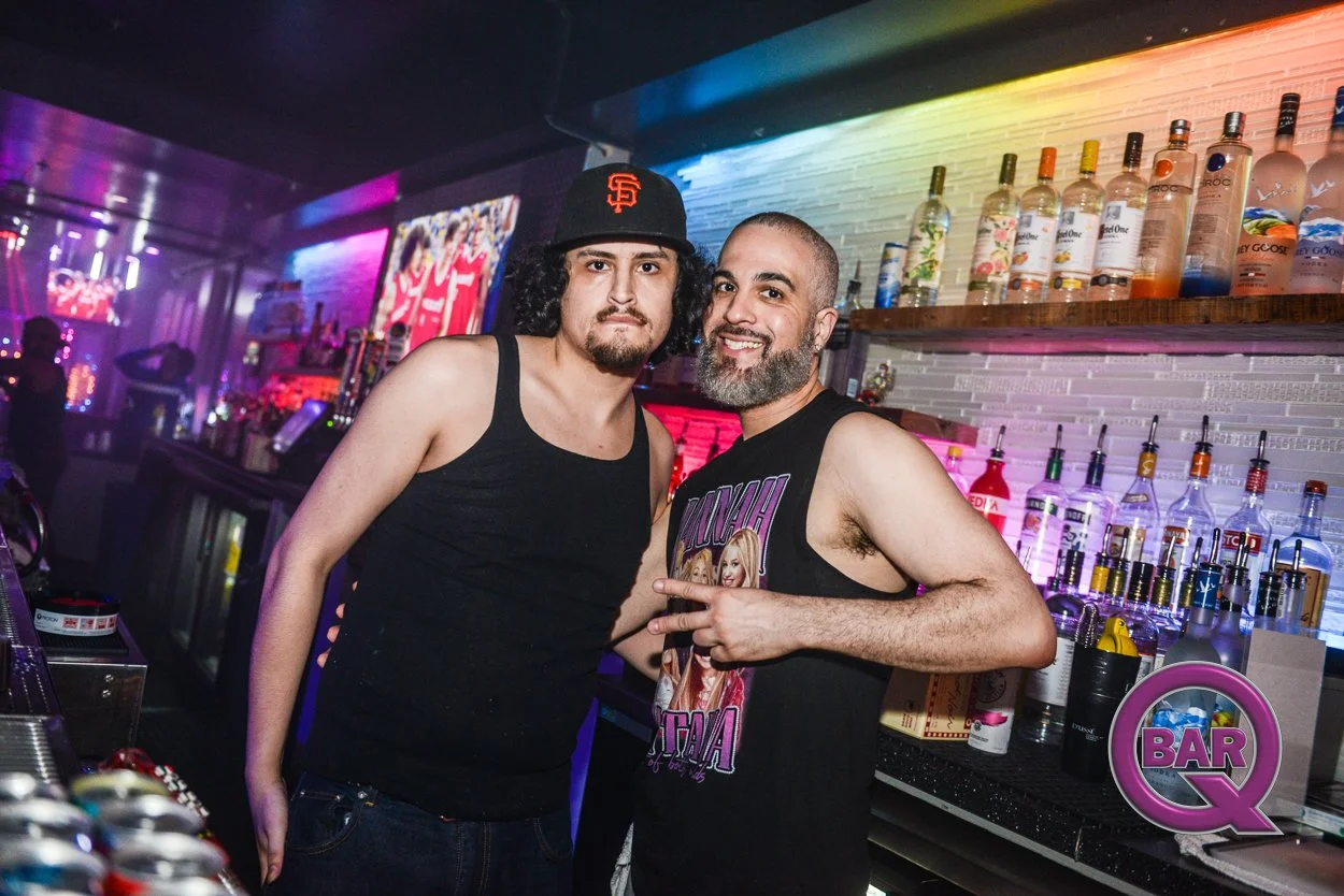 Two men standing at a bar, one with curly black hair wearing a black tank top and a San Francisco Giants cap, the other with a beard and shaved head wearing a sleeveless graphic T-shirt showing the Spice Girls, smiling and making a peace sign. The ba