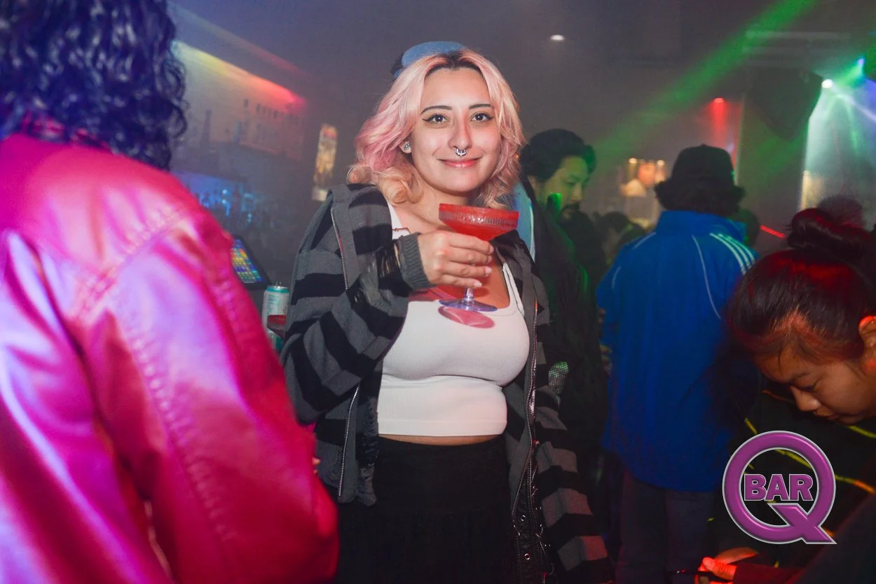A woman with pink hair and a septum piercing holding a red cocktail in a bar or nightclub, surrounded by other people, with colorful lights.