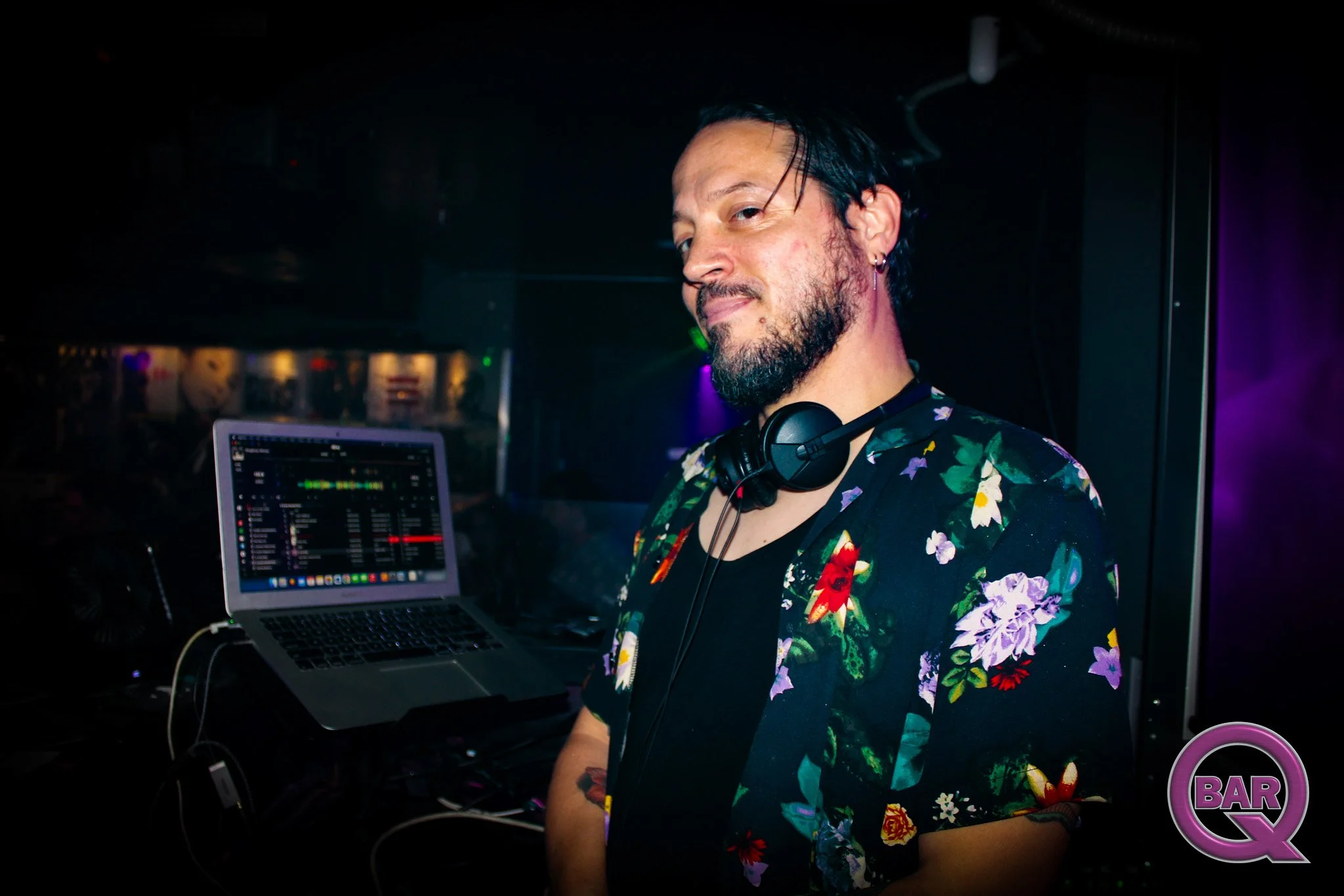 A male DJ with a beard and earrings wearing a colorful floral shirt, standing in a dimly lit club with a laptop and DJ equipment, smiling slightly.