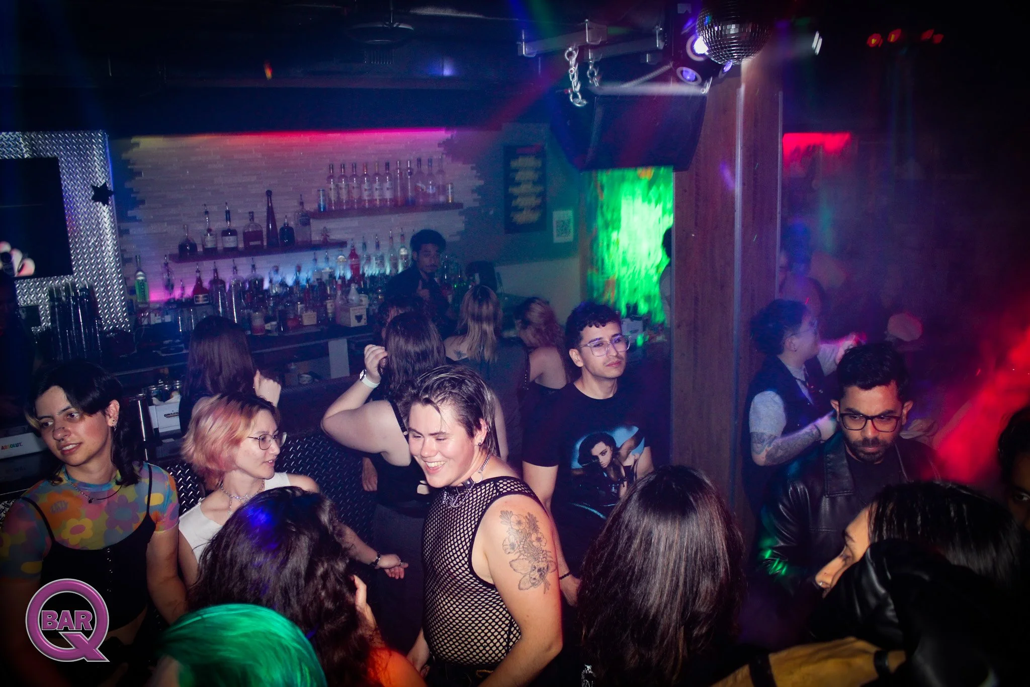 A crowded nightclub with people dancing and socializing under colorful lights, a visible bar with bottles in the background, and a disco ball hanging from the ceiling.