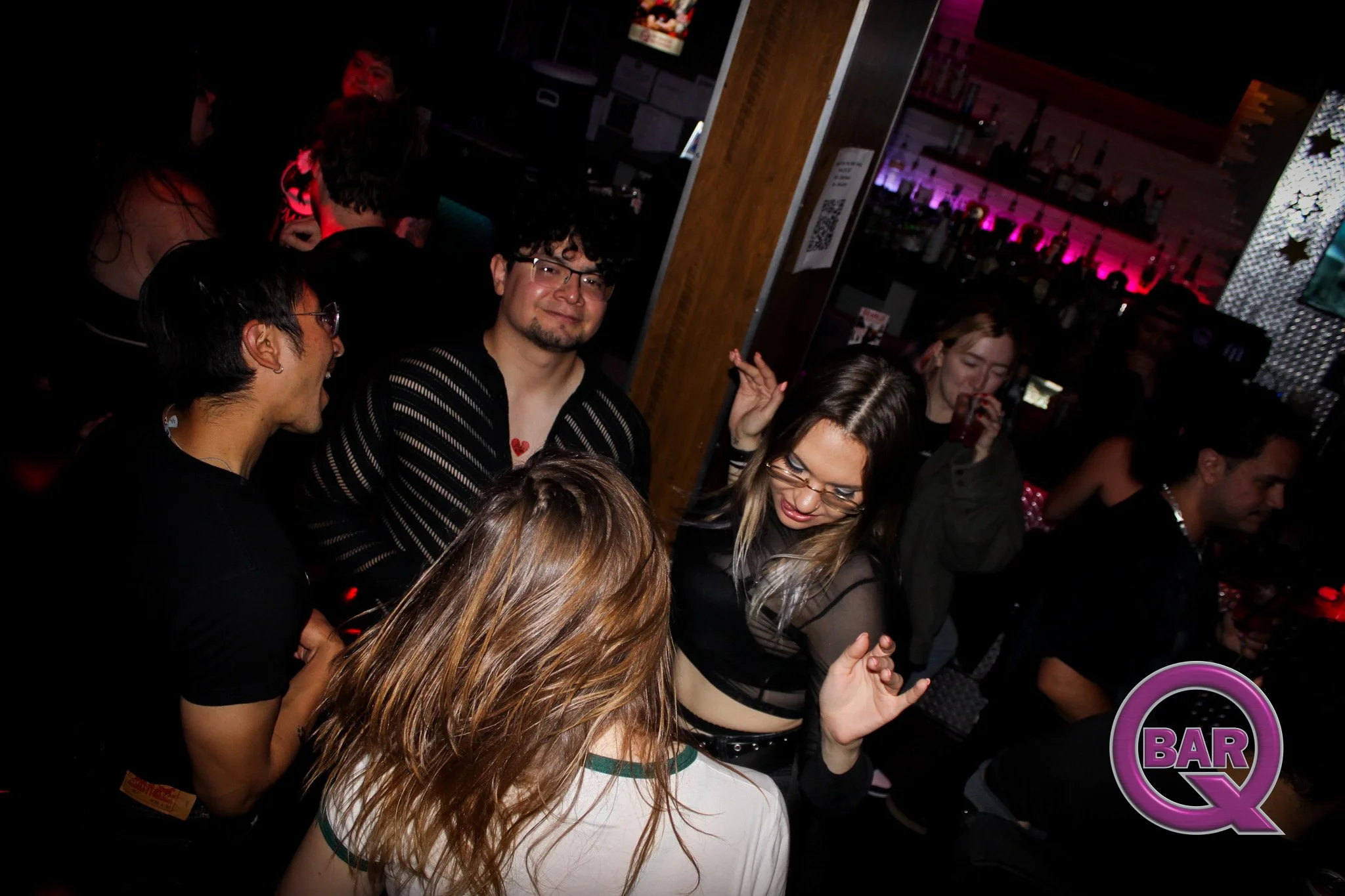 A group of young people socializing at a bar or nightclub with dim lighting, some women are drinking, and some men are talking, with shelves of bottles visible in the background.
