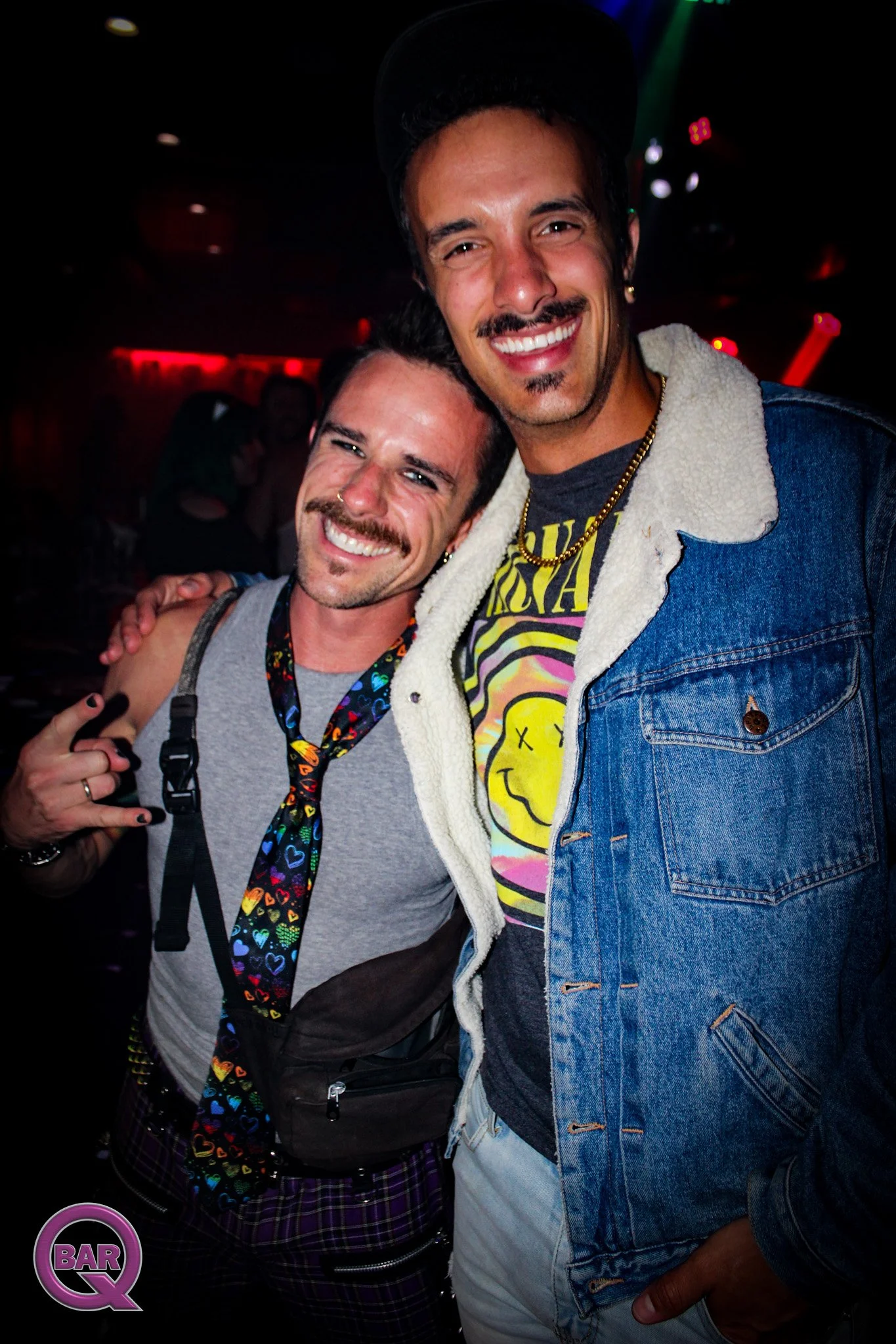 Two smiling men in a dark indoor setting, one wearing a denim jacket with a colorful graphic t-shirt, and the other wearing a gray tank top with a rainbow heart-patterned lanyard, both showing happy expressions.