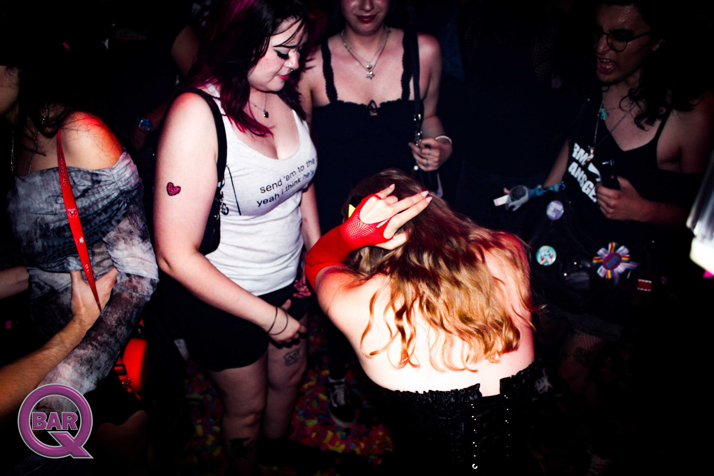 A group of people gathered closely at a party or nightclub, some with colorful hair and wearing dark clothing, with some holding drinks. The scene is dimly lit, and one person is touching their head while others look on.