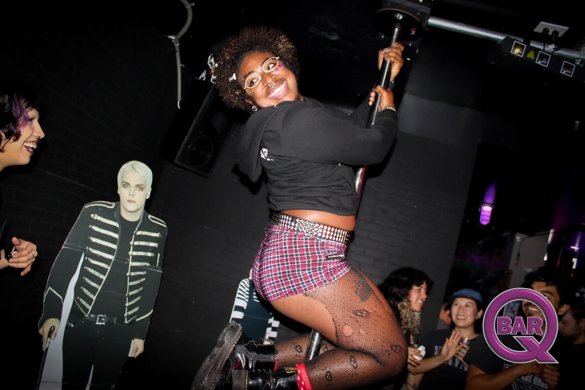 A person with curly hair and glasses riding a bicycle at a bar or nightclub, surrounded by people and bar signs.