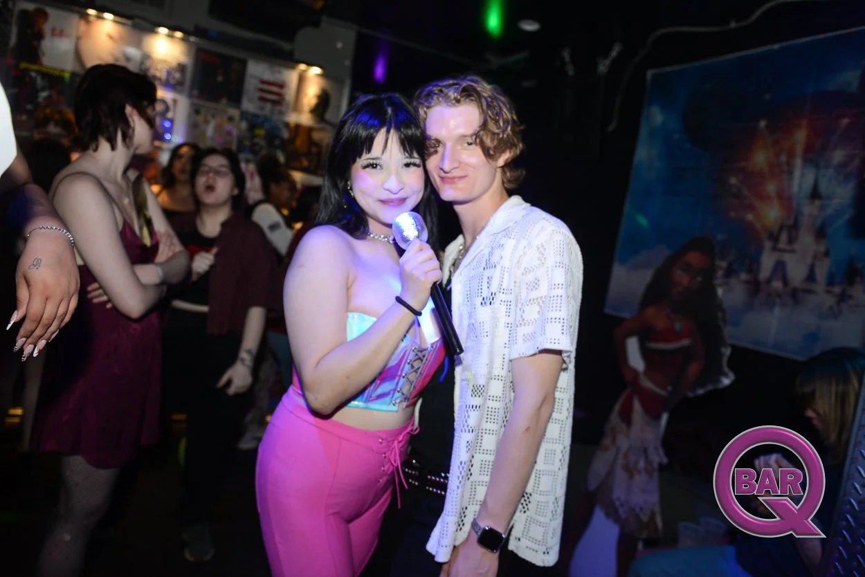 A woman in a colorful top and pink pants holding a microphone, standing beside a man in a white patterned shirt inside a lively bar with other people in the background.