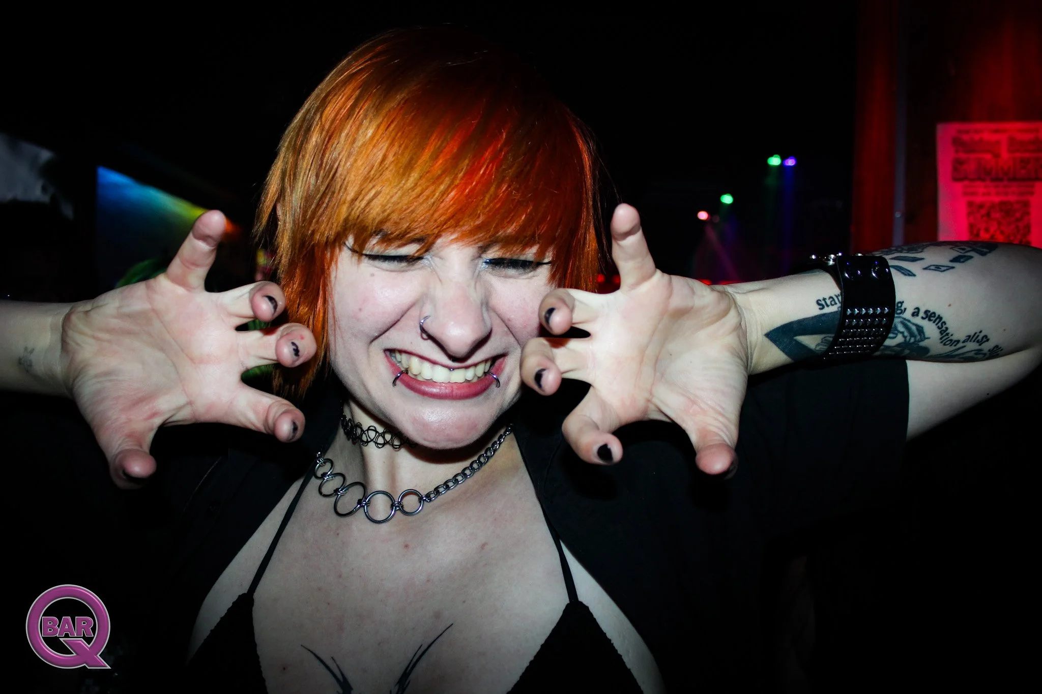 Person with orange hair smiling and holding hands near face, in a dark setting with colorful lights in the background.