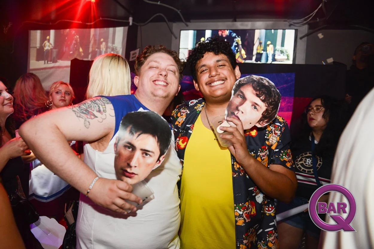 Two men smiling and holding large cutouts of celebrity faces at a lively bar or club with other people in the background and a screen displaying visuals.