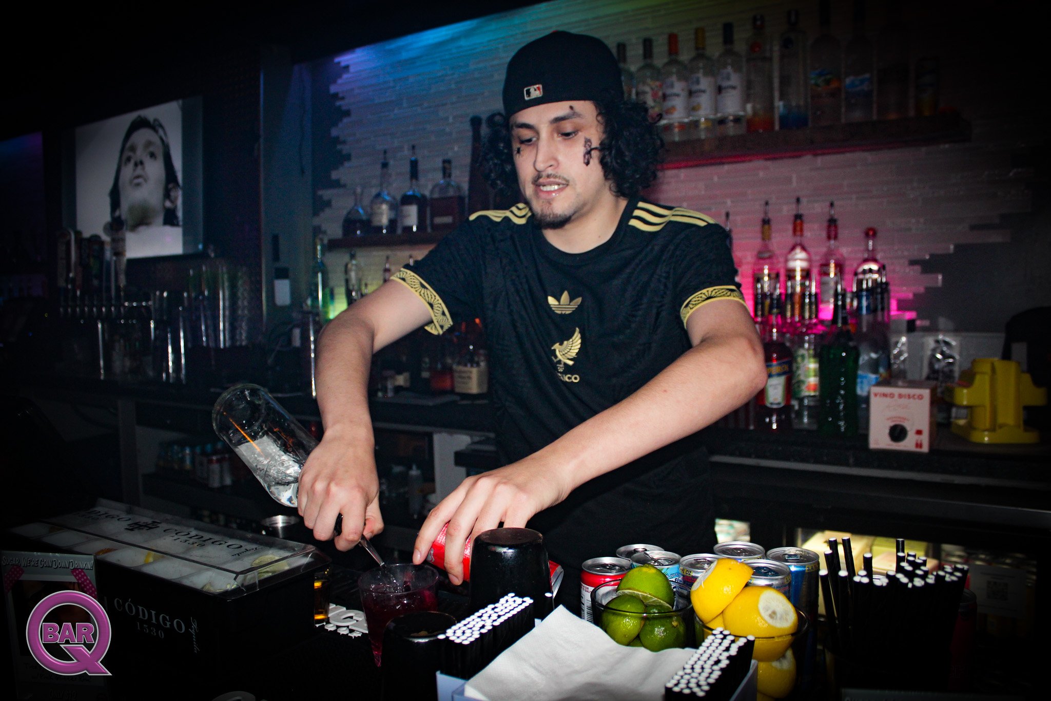 A man with curly black hair, tattoos on his face, wearing a black baseball cap and a black Adidas shirt, preparing drinks at a bar. The bar has various bottles and cans, limes, lemons, and mixers on the counter.
