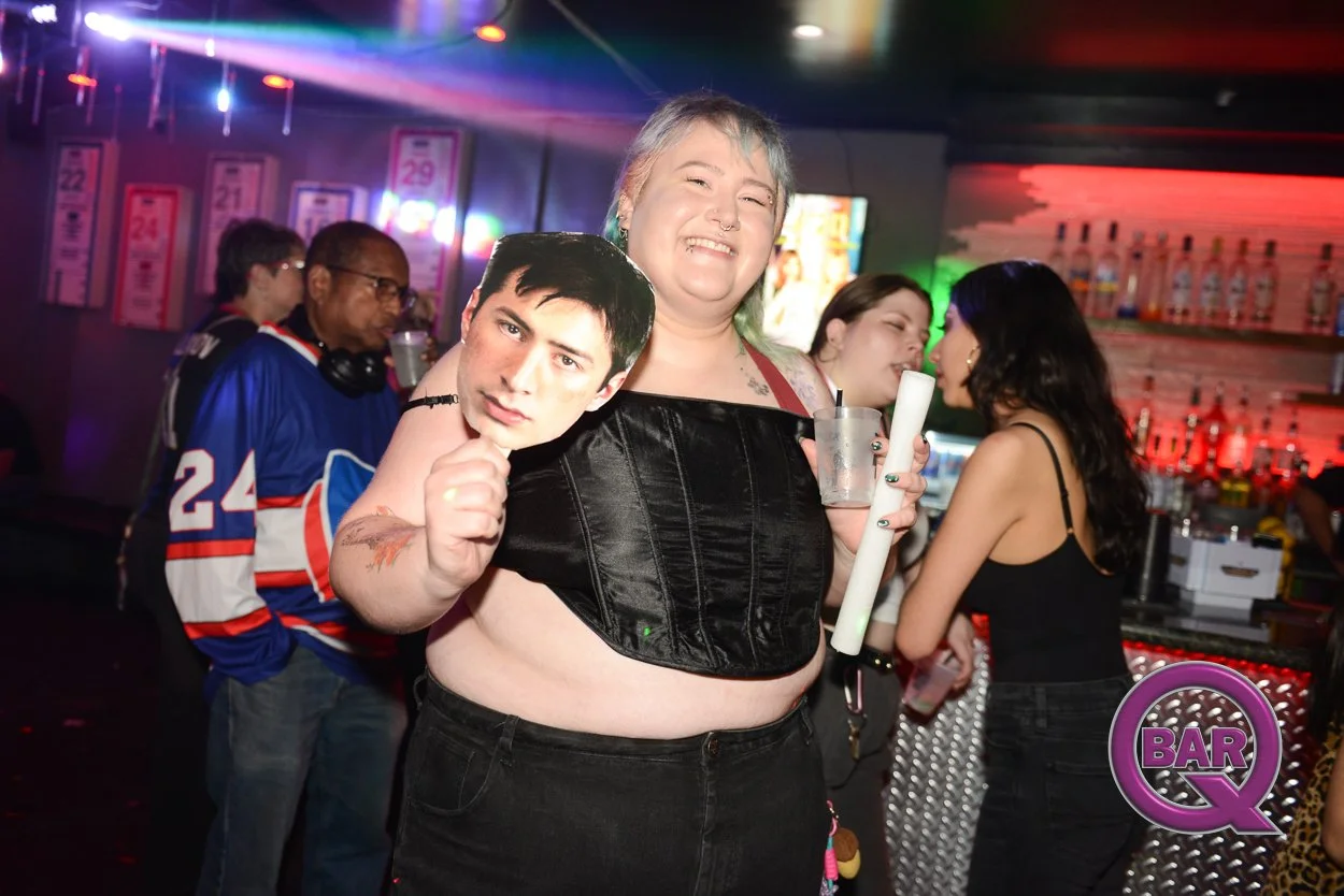 A smiling person at a bar holding a large cutout of a man's face, with other people and bar signage in the background.