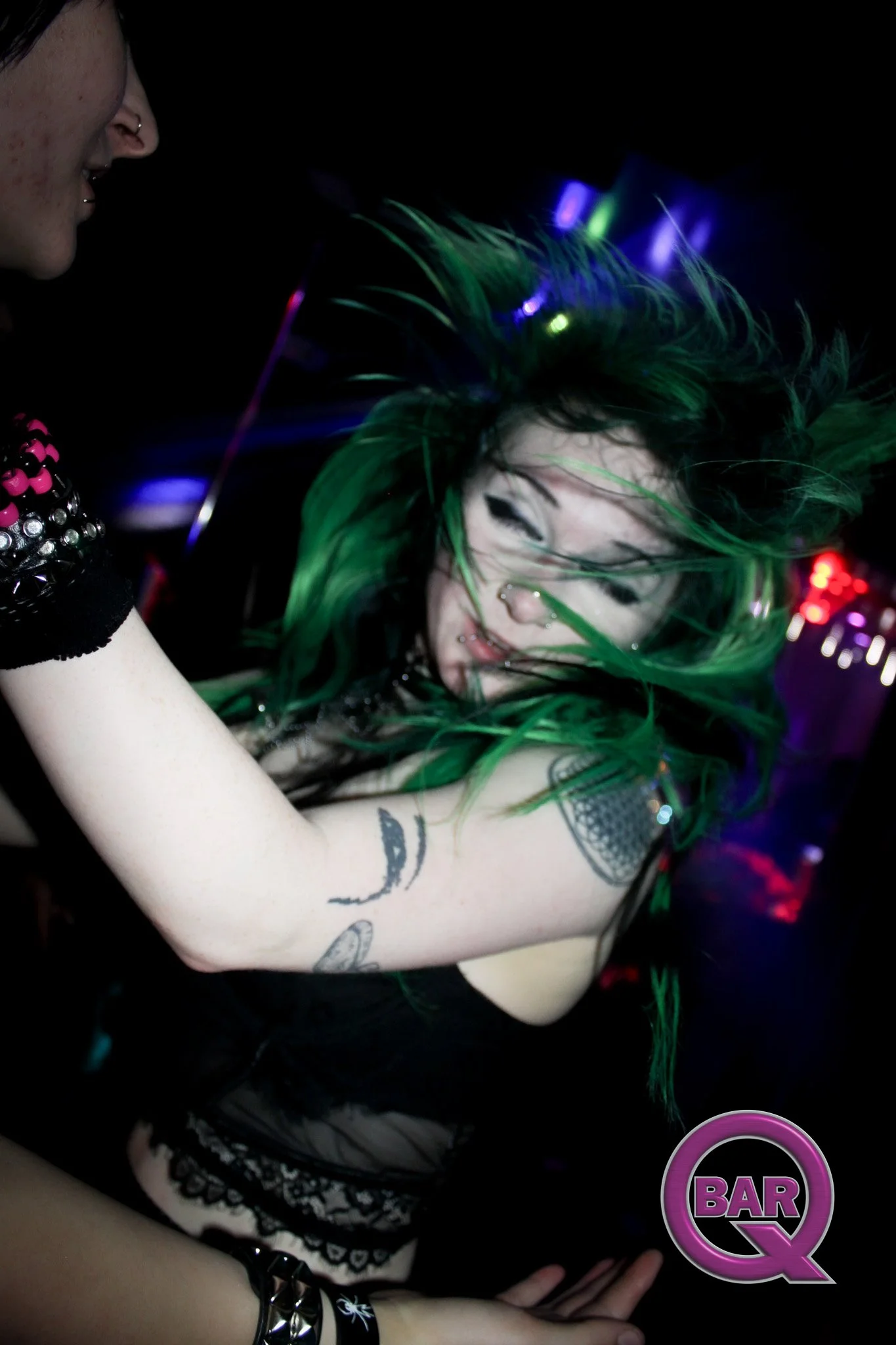 A woman with vibrant green hair and tattoos on her arm, wearing black lace clothing and bracelets, is in a dark room with colorful lights. She appears to be dancing or moving to music, with a cheerful expression. There is a pink and purple 'BAR Q' lo