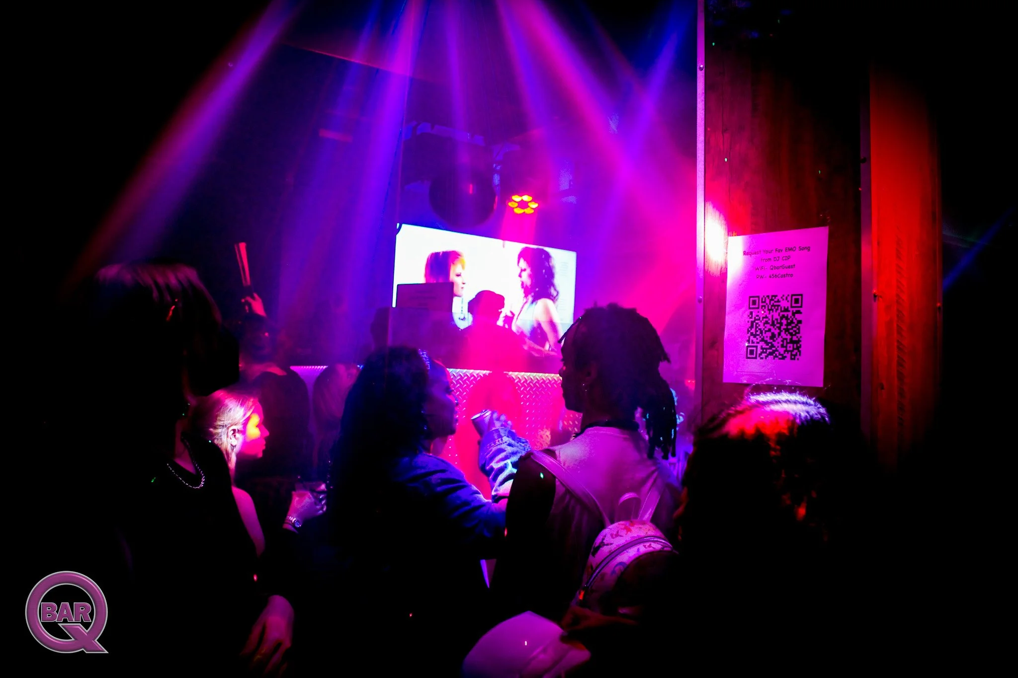 Dimly lit nightclub with purple and pink stage lights, showing a large screen with two women speaking. Visible audience members are facing the stage, with some holding drinks and taking photos.
