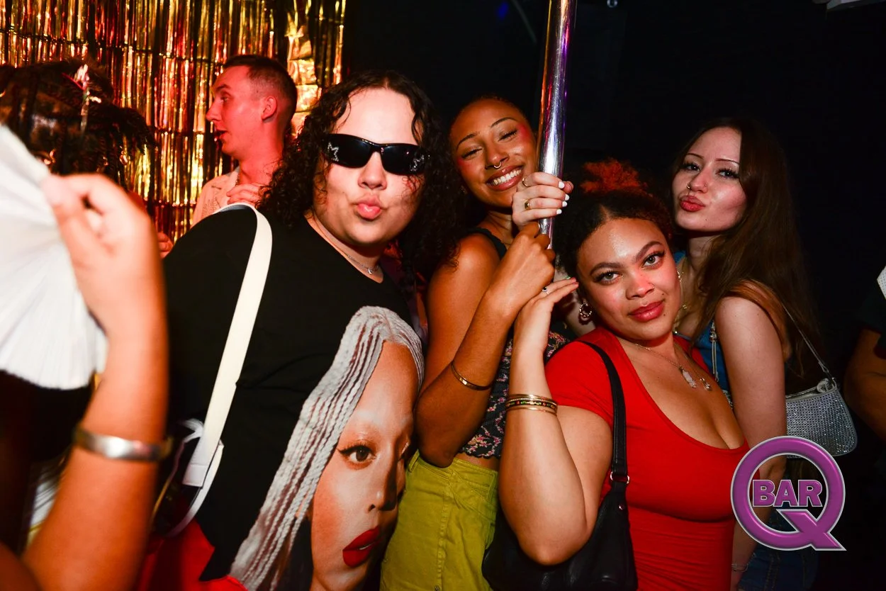 Group of five women and one man posing at a bar or club with colorful lighting and a gold streamers backdrop.
