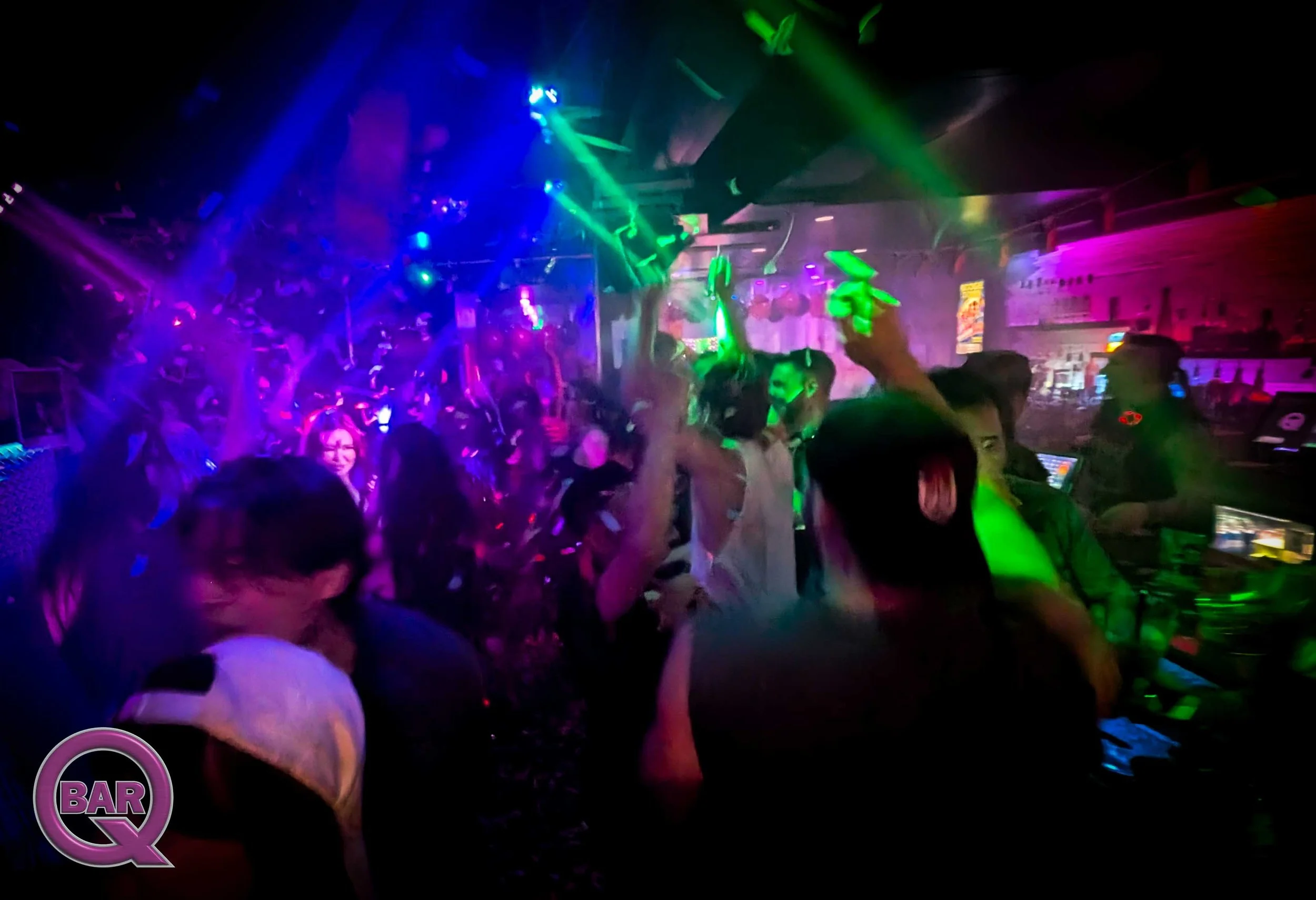People dancing in a club with colorful lights, a bar area in the background, and a DJ booth.