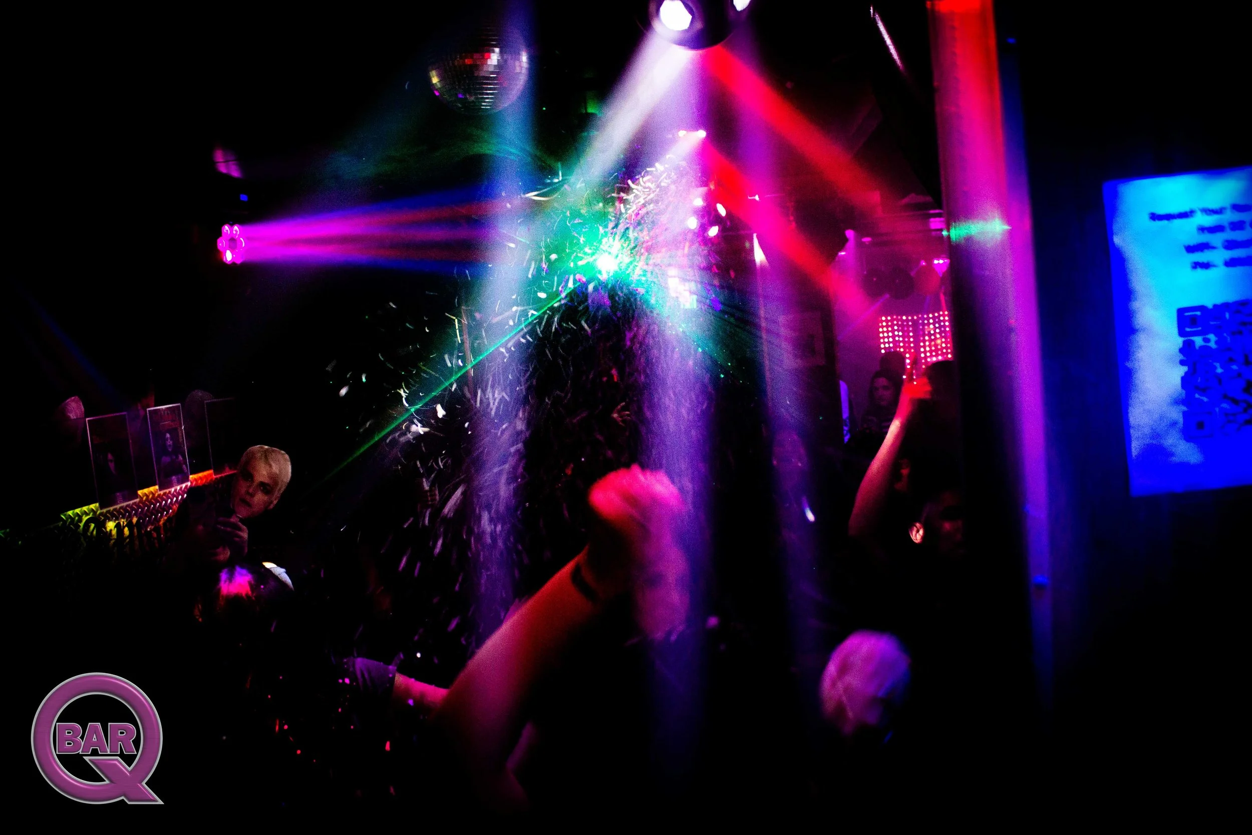 Dance floor scene at a nightclub with colorful lighting, a disco ball, and partygoers dancing. Purple and pink lights illuminate the scene, and there's confetti in the air. A person wearing a red shirt is visible on the right, and a woman with blonde