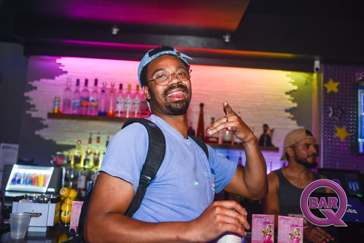 A man with glasses and a backpack making a peace sign inside a bar with colorful lights and bottles in the background.