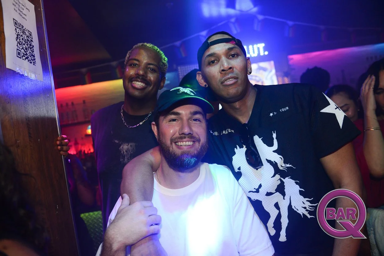 Three men smiling and posing together in a nightlife setting with colorful lighting. The man in the center wears a white shirt and a black cap, while the men on either side wear dark shirts; one has a cap and sunglasses hanging from his shirt. There 