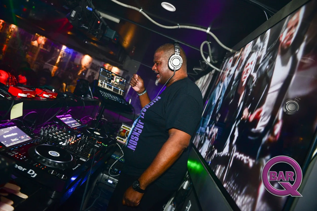 A DJ in a nightclub or bar mixes music on a DJ controller with a laptop and sound equipment. The DJ is wearing headphones and a black t-shirt, dancing and enjoying himself. There are colorful lights and a large screen behind him displaying visuals, w