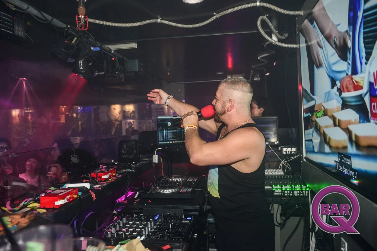 A DJ in a black tank top with a red microphone and a tattooed arm performs in a nightclub, surrounded by DJ equipment and a crowd of people dancing.