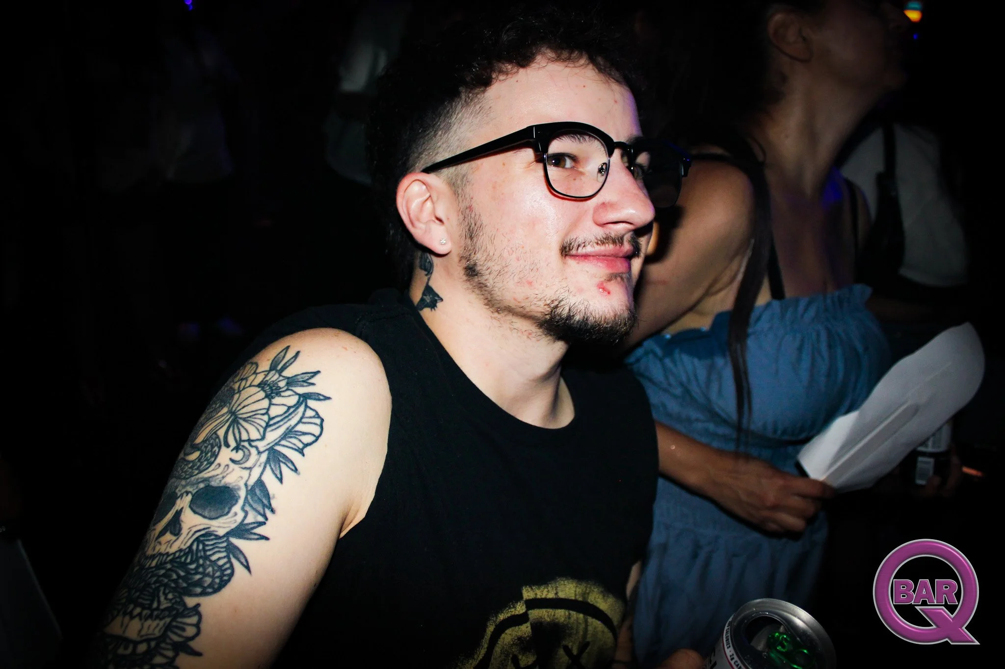 A young man with short dark hair and glasses, showing tattoos on his arm and neck, smiling in a dark setting with other people around. He is holding a drink and appears to be at a social event.