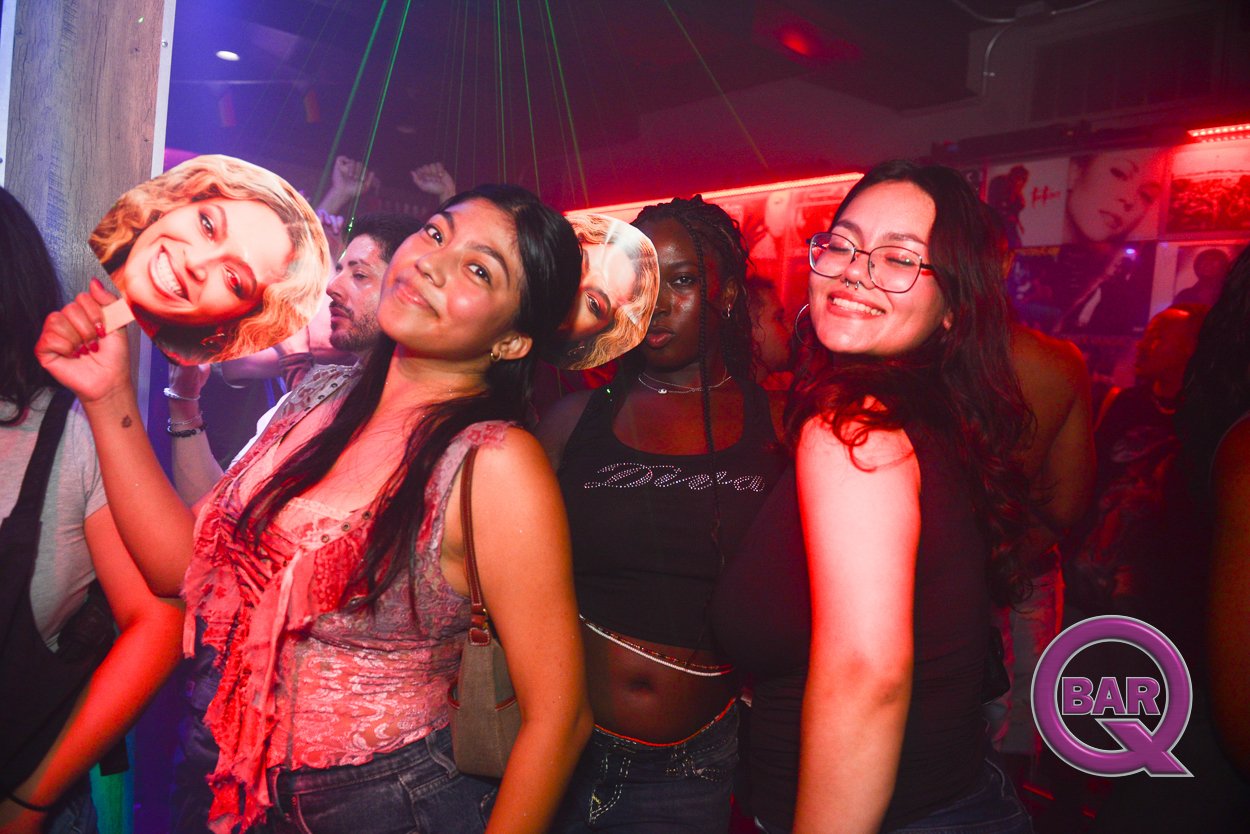 People dancing and having fun at a nightclub, with a woman holding a cardboard cutout of a smiling woman's face.