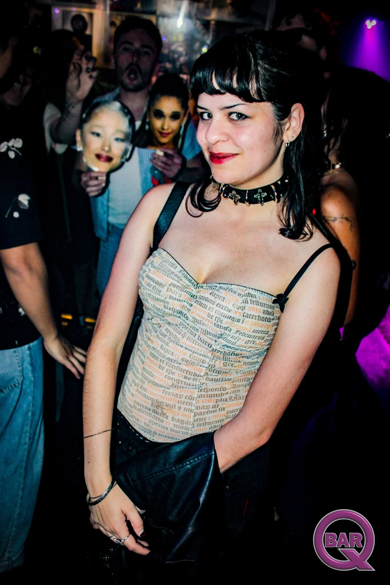 Close-up of a woman with dark hair, wearing a necklace with crosses, taking a selfie at a crowded bar or club. In the background, there are people, some holding masks, and purple lights.
