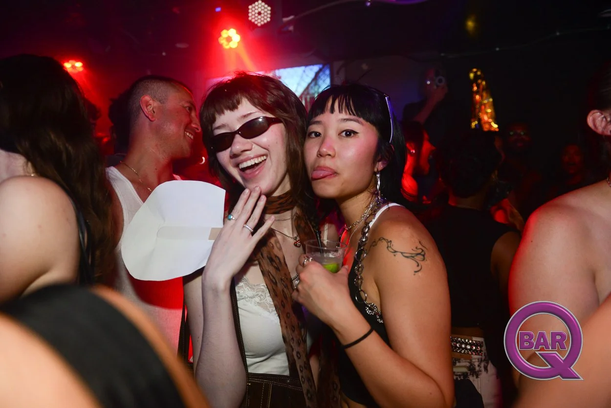 Two women at a party or club, one wearing sunglasses and laughing, the other sticking out her tongue, surrounded by people in a lively, colorful setting.