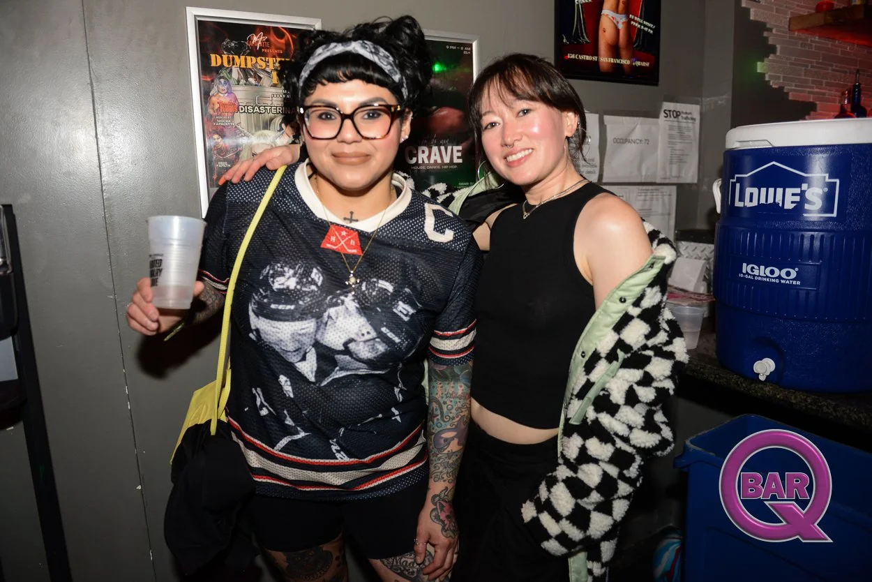 Two women smiling and posing together at a bar, one wearing glasses and a sports jersey with tattoos, the other wearing a black top and a black and white checkered hoodie, behind a counter with a blue Lowe's ICLOO water cooler.