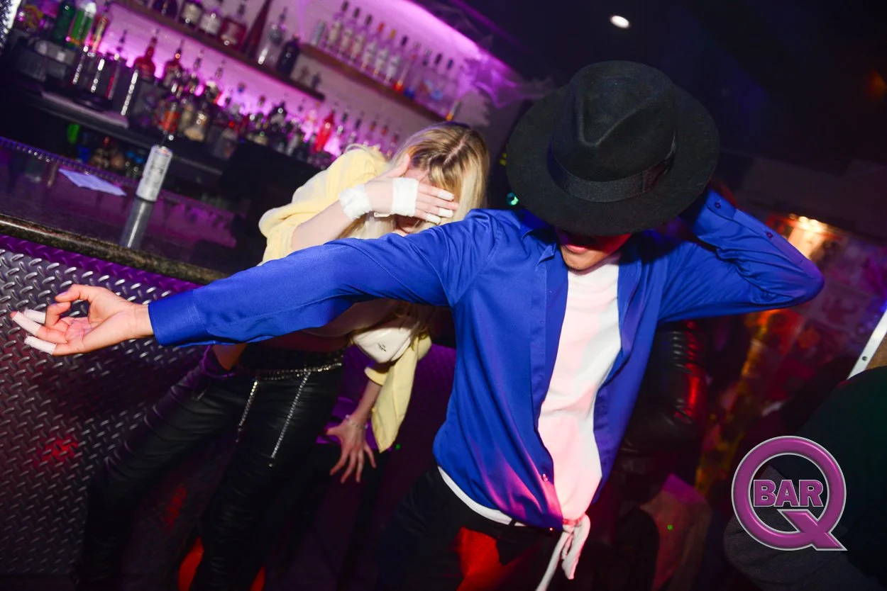 A person in a blue shirt and black hat dancing with artwork on their face obscuring their features, next to a woman with blonde hair and a yellow jacket, in a bar with purple lighting and bottles on shelves.