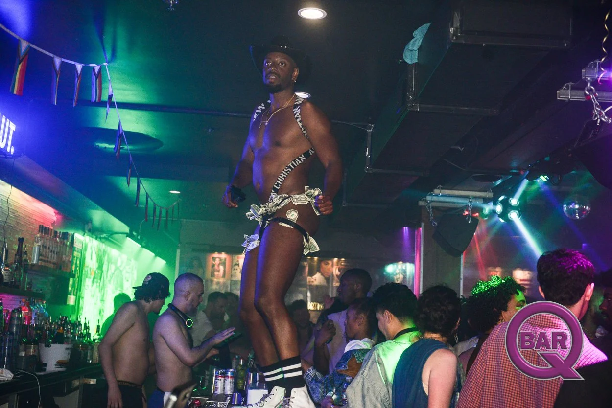 A shirtless man dancing on a bar counter in a nightclub with colorful lighting and a crowd of people around him.
