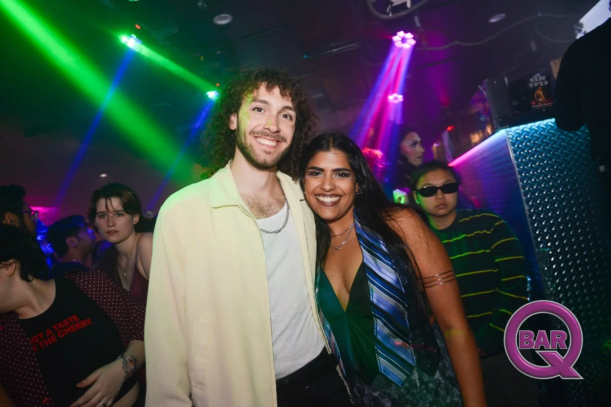 People dancing and socializing in a nightclub with colorful lights, including a woman and a man smiling at the camera in the foreground. There is a woman with sunglasses and a striped shirt in the background, along with other party-goers.