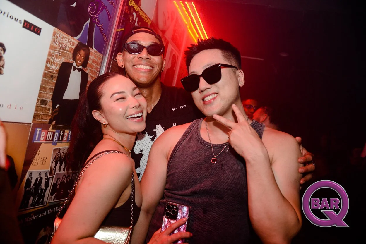 Three people smiling and enjoying themselves at a nightclub, wearing sunglasses, with posters on the wall and red lighting.