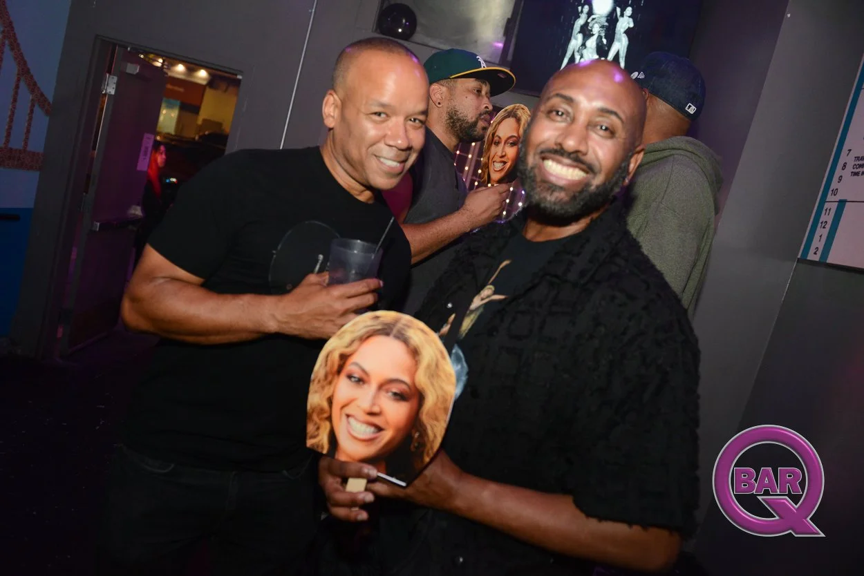 Two men smiling and holding a cutout photo of a woman's face at a bar or club. One man is wearing a black shirt and the other a textured black shirt. People are in the background, including a person in a gray hoodie and another in a baseball cap, wit