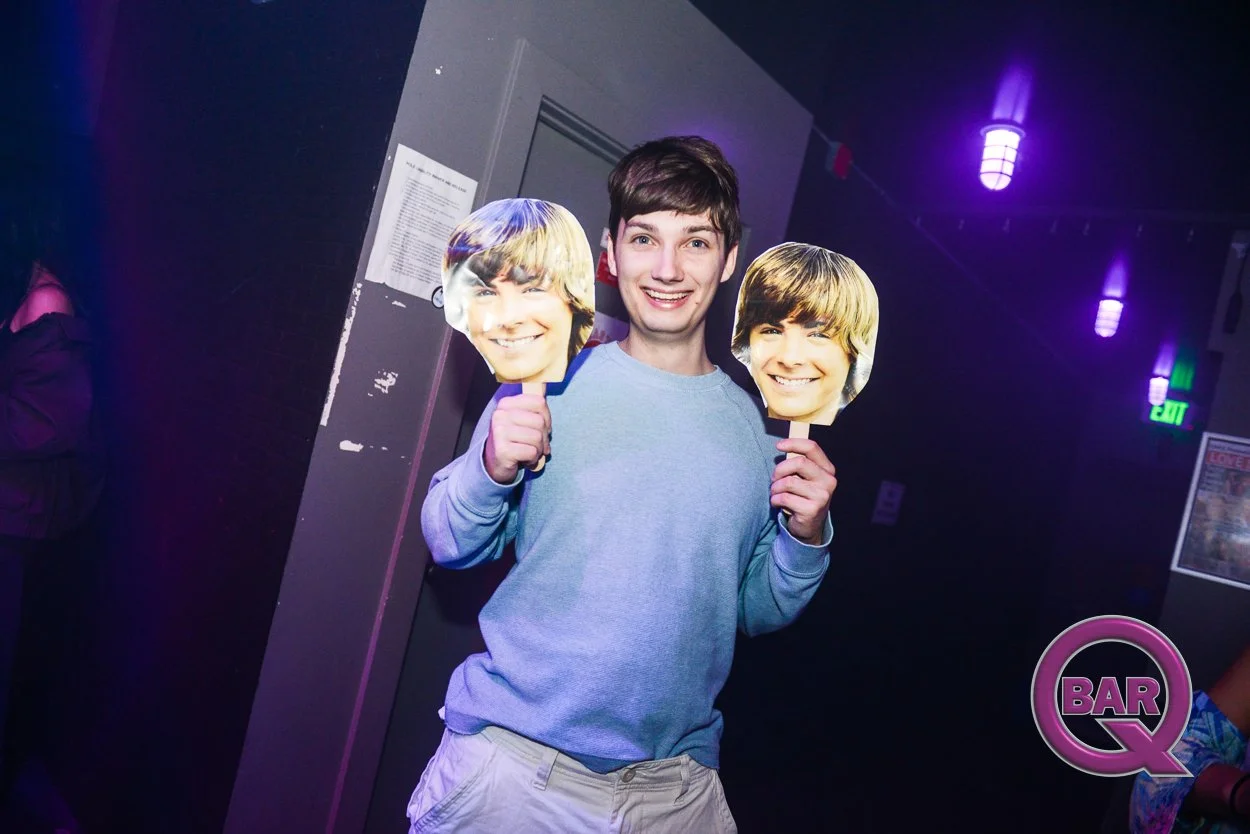 A young man holding two fans with images of a young boy's face, smiling at a bar or nightclub with purple lighting and an exit sign in the background.