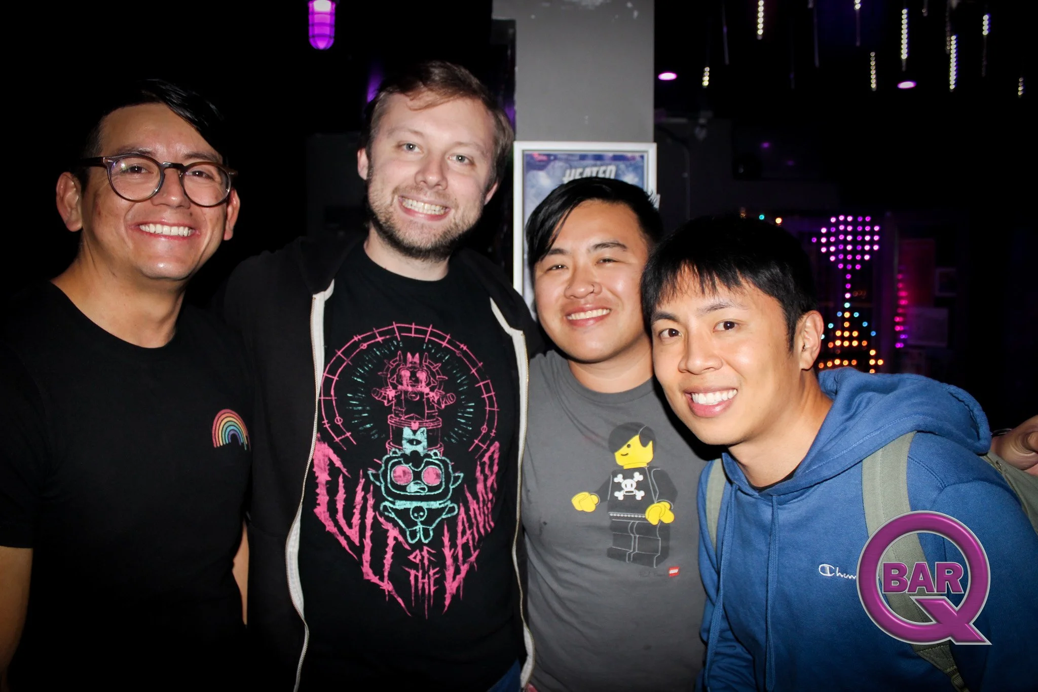 Four smiling men standing close together inside a bar or nightclub with colorful neon lights in the background.